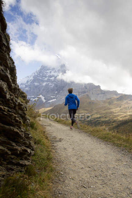 Vista trasera del hombre corriendo por la pista de tierra hacia el monte Eiger, Grindelwald, Suiza - foto de stock