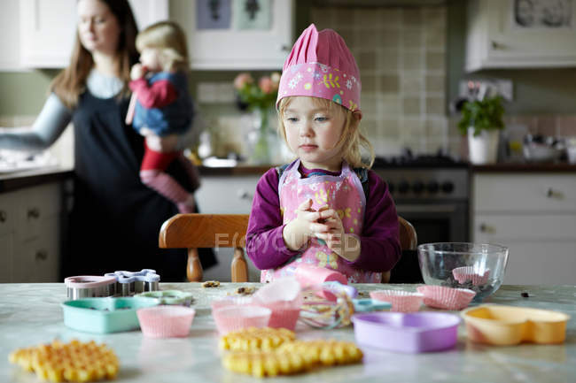 Toddler girl baking in kitchen, mother with child in background — Stock Photo