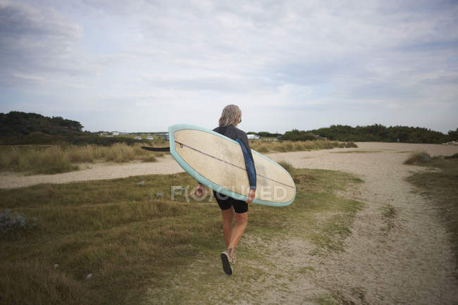 Senior woman walking along beach, carrying surfboard, rear view — Stock Photo