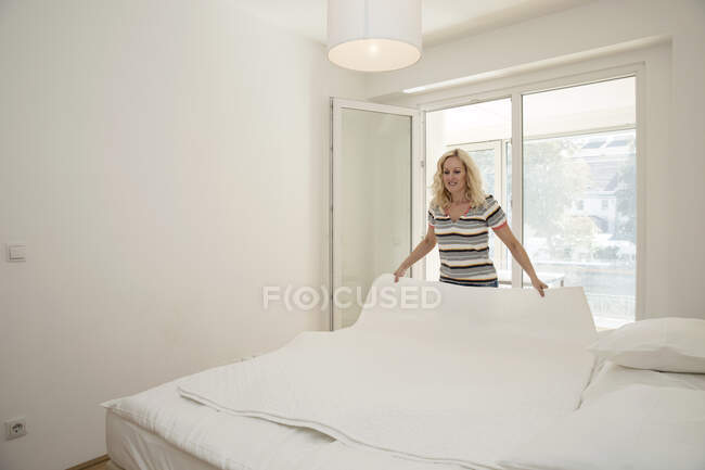 Зрелая женщина в спальне держит одеяло застилающее постель — стоковое фото