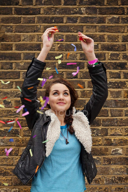 Mujer joven cayendo confeti colorido contra la pared de ladrillo - foto de stock