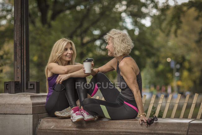Dos amigas maduras entrenando en el parque, sentadas en la pared con café para llevar - foto de stock