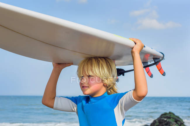 Молодий хлопець на пляжі, носить дошку для серфінгу на голові — стокове фото