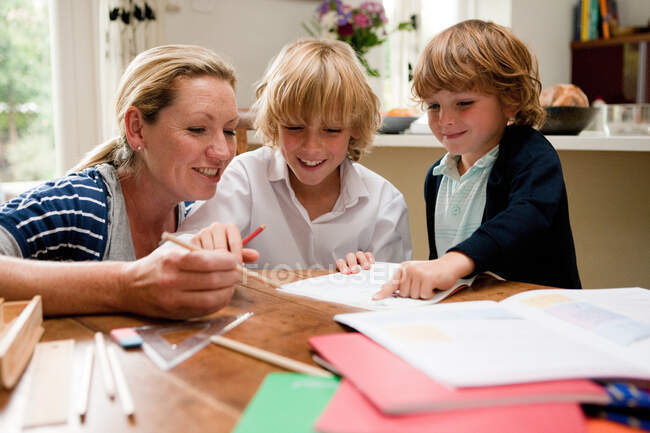 Мама допомагає старшому синові виконувати домашні завдання за столом. — стокове фото