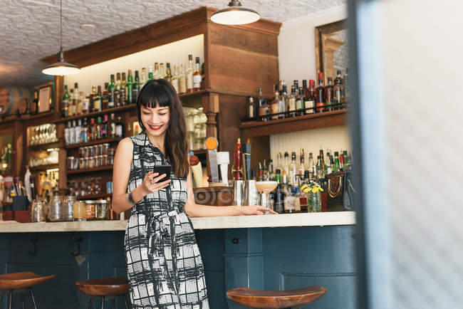 Mujer joven leyendo texto del teléfono inteligente en el bar de cócteles - foto de stock