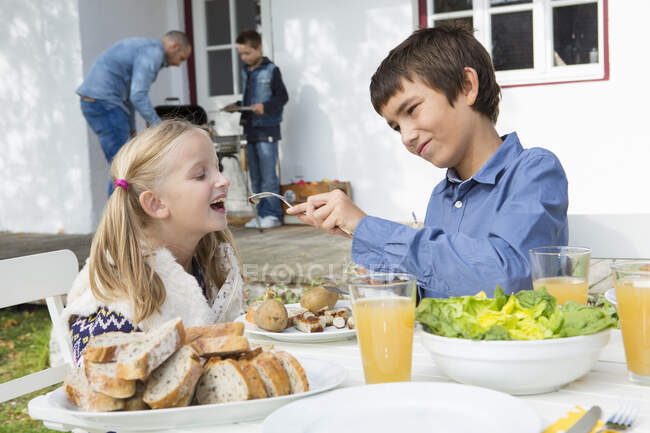 Підліток хлопчик годує сестру в саду барбекю столом — стокове фото