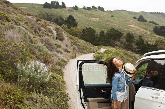 Young woman standing by car on country road, smiling — Stock Photo