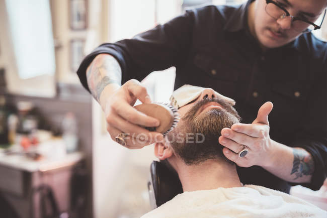 Barbeiro escovando barba cliente na barbearia — Fotografia de Stock
