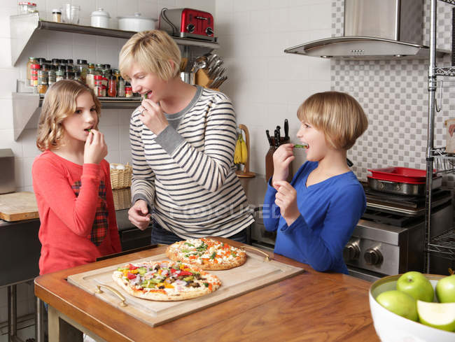 Madre y dos hijas en la cocina preparando comida, mordisqueando los ingredientes - foto de stock
