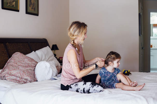 Woman sitting on bed plaiting toddler daughter hair — Stock Photo