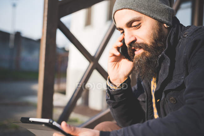 Mittlerer erwachsener Mann, im Freien sitzend, mit Handy und digitalem Tablet — Stockfoto