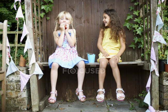 Retrato de dos chicas sentadas en el banco comiendo un cubo de fresas frescas - foto de stock