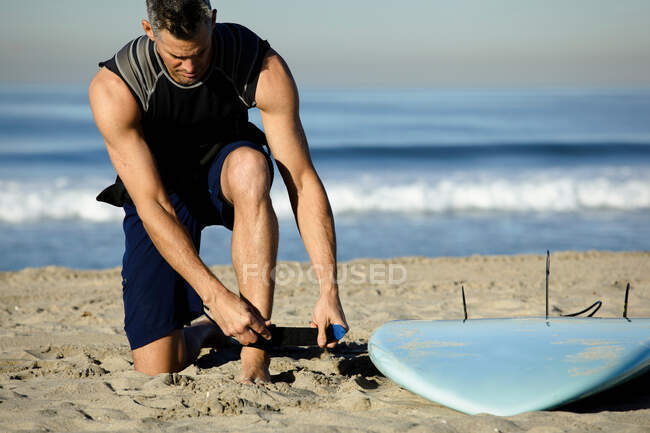 Средний взрослый мужчина пристегивает ремень на пляже для серфинга — стоковое фото