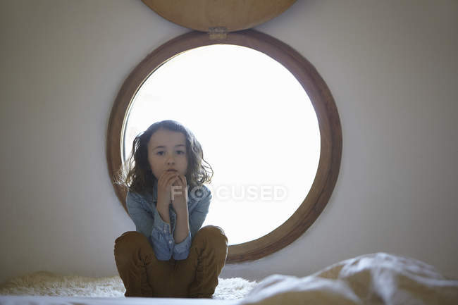Porträt eines mürrischen Mädchens, das vor einem kreisrunden Fenster hockt — Stockfoto