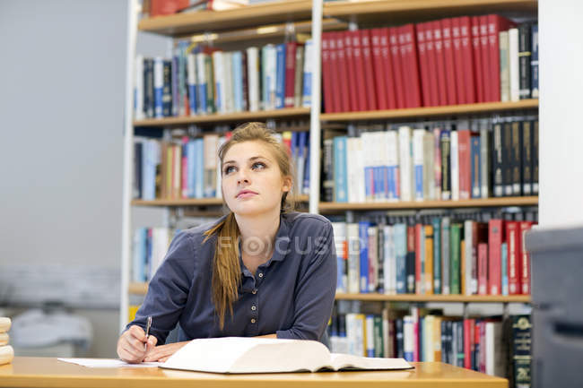 Young female student reading textbook gazing up from library desk — Stock Photo