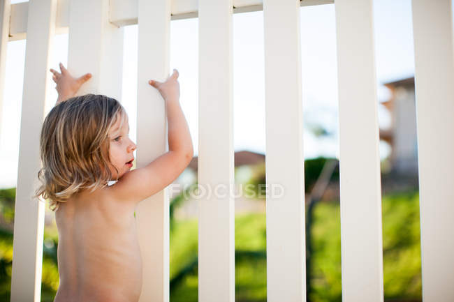 Young boy outdoors, holding onto white fence — Stock Photo
