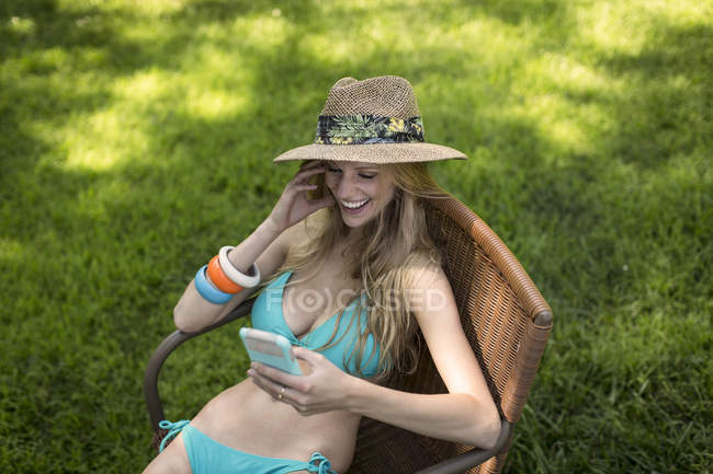 Mujer joven con bikini y sombrero de sol leyendo textos de teléfonos inteligentes en el jardín - foto de stock
