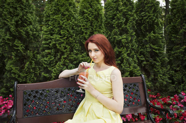 Mujer joven bebiendo jugo en el banco del parque - foto de stock