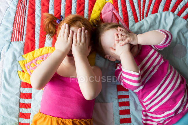 Retrato de dos hermanas jóvenes acostadas sobre una manta, cubriendo la cara con las manos - foto de stock