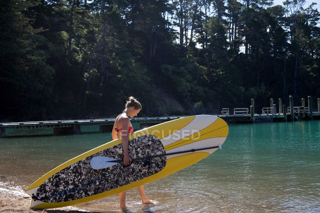Mulher adulta média carregando paddleboard no mar — Fotografia de Stock