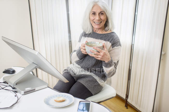 Portrait de la femme âgée à la maison travaillant à son ordinateur de bureau à domicile prenant du café / thé — Photo de stock