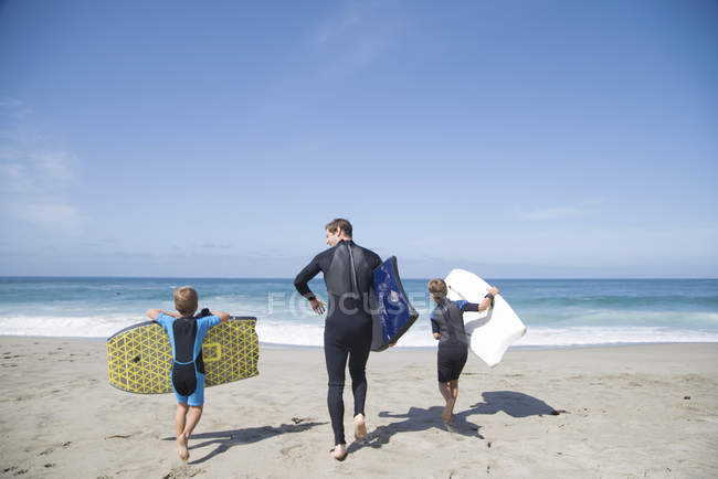 Rear view of man and two sons running down beach and carrying bodyboards, Laguna Beach, California, USA — Stock Photo
