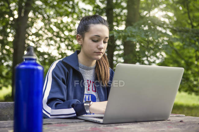 Дівчина-підліток в парку використовує ноутбук на лавці для пікніка — стокове фото