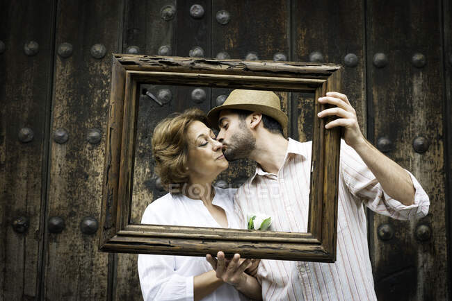 Portrait of adult son kissing mother on cheek, holding wooden frame in front of their faces, Mexico City, Mexico — Stock Photo