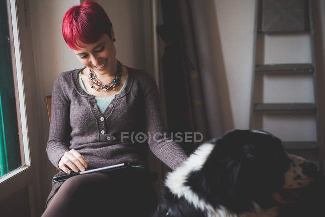Молода жінка вдома використовує цифровий планшет, погладжуючи собаку — стокове фото