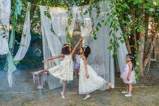 Dos chicas jóvenes, vestidas de hadas, bailando al aire libre, una chica más joven mirando desde el costado - foto de stock