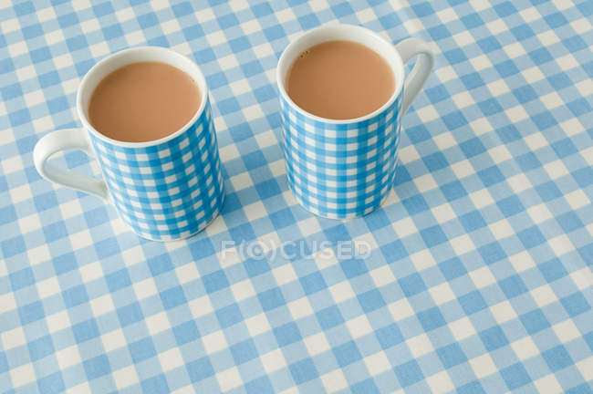 Vista ravvicinata di due tazze di tè con latte sullo sfondo del motivo a quadretti — Foto stock