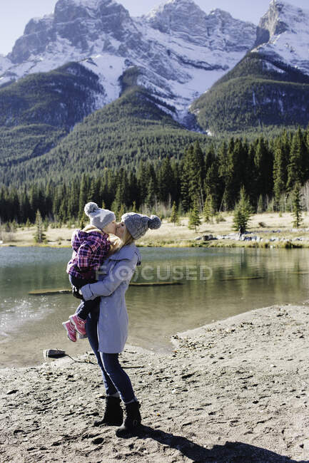 Madre sosteniendo y besando hija junto al río, Tres hermanas, Montañas Rocosas, Canmore, Alberta, Canadá - foto de stock