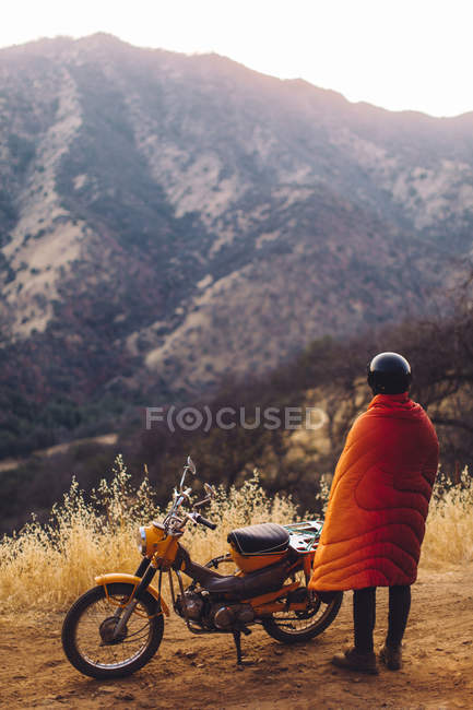 Hombre de pie junto a la moto, envuelto en manta, mirando a la vista en el Parque Nacional Sequoia, California, EE.UU. - foto de stock