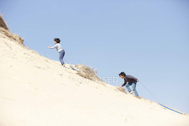 Deux jeunes garçons escaladant une colline sablonneuse, tirant des traîneaux derrière eux — Photo de stock