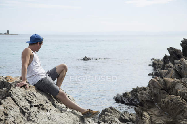 Вид сбоку в полный рост молодого человека, сидящего на скалах и глядящего в сторону океана, Стинтино, Италия — стоковое фото
