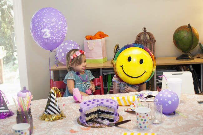 Duas meninas sentadas na mesa de festa de aniversário com bolo brincando com balão de rosto sorridente — Fotografia de Stock