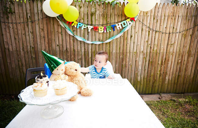 Bébé garçon assis à table avec des jouets doux portant un chapeau de fête — Photo de stock