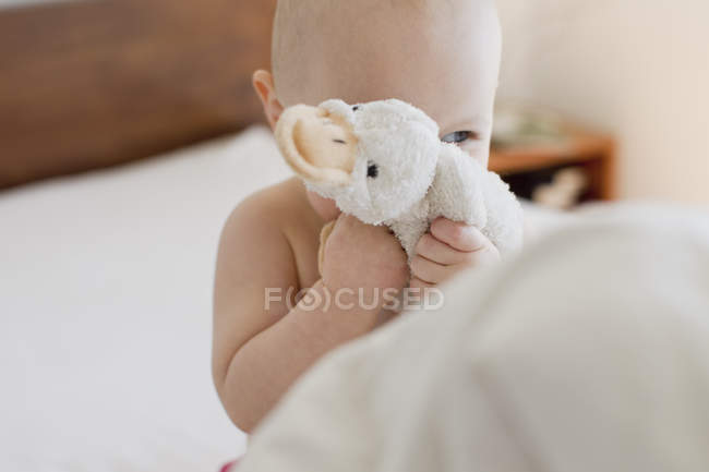 Baby girl sitting up in bed hiding behind soft toy — Stock Photo