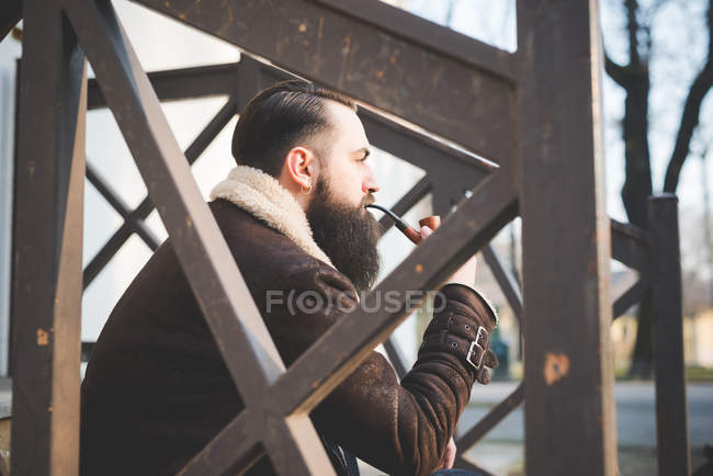 Молодой бородатый мужчина курит трубку на ступеньках — стоковое фото