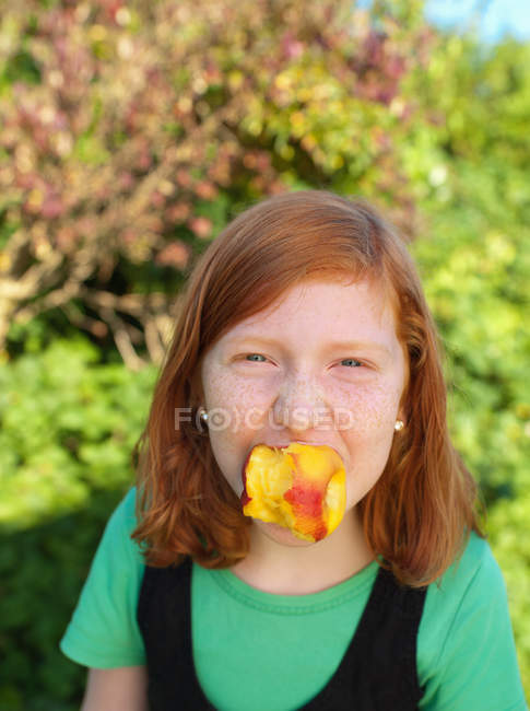 Young girl holding peach in mouth, portrait — Stock Photo