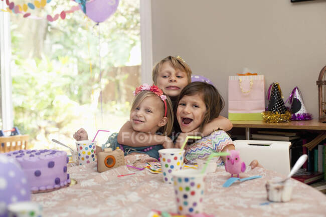 Three children sitting at birthday party table hugging each other — Stock Photo