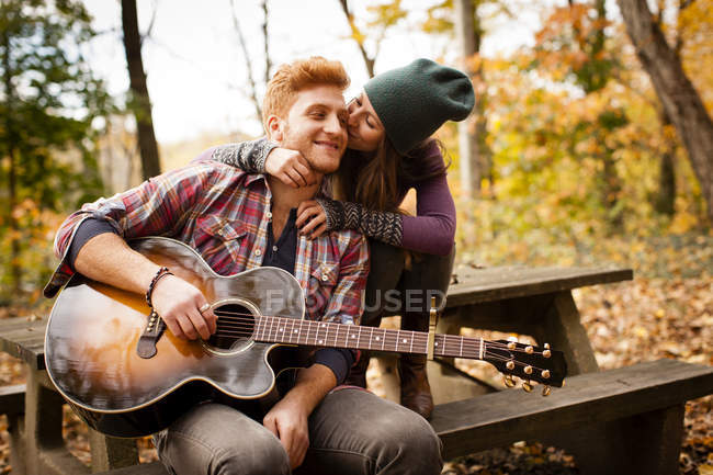 Romántica pareja joven tocando la guitarra en el banco de picnic en el bosque de otoño - foto de stock