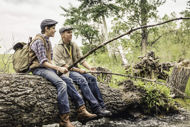 Uomo e ragazzo che indossano berretti piatti seduti su un albero caduto attraverso la pesca fluviale con rami — Foto stock