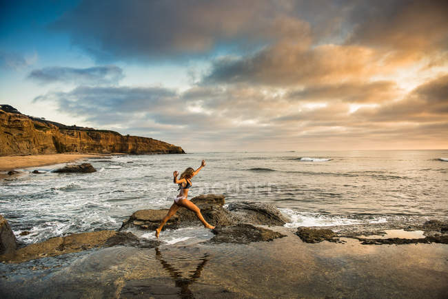 Young woman in bikini leaping from rocks on beach — Stock Photo
