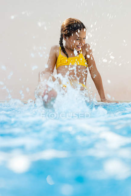 Chica sentada a la orilla de la piscina pateando y salpicando - foto de stock