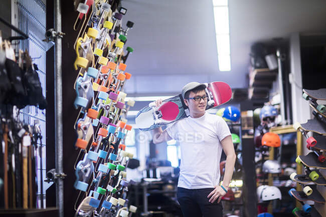Молодий чоловік скейтбордист зі скейтбордом над плечем в магазині скейтбордів — стокове фото