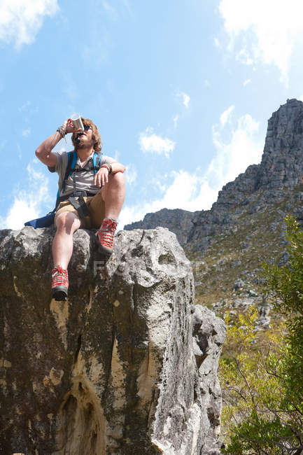Männlicher Wanderer sitzt auf Felsformation und trinkt aus Flasche — Stockfoto