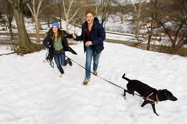 Casal jovem correndo com cão no parque central nevado, Nova York, EUA — Fotografia de Stock