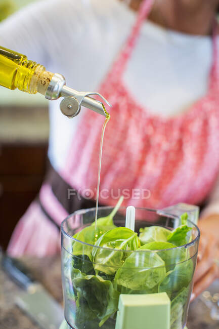Donna matura versando olio d'oliva nel robot da cucina contenente basilico, facendo salsa di pesto, sezione centrale — Foto stock