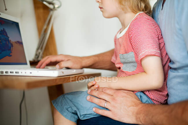 Boy sitting on father's lap at computer desk — Stock Photo